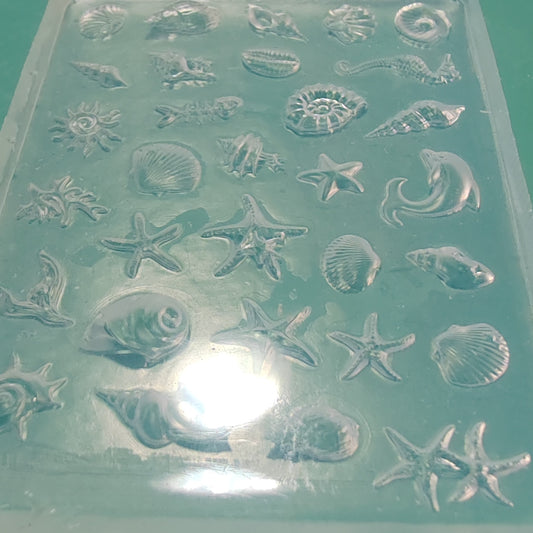 A translucent silicone mold tray featuring embossed sea-themed designs including starfish, seashells, dolphins, and a spiral shell, shown on a green background.