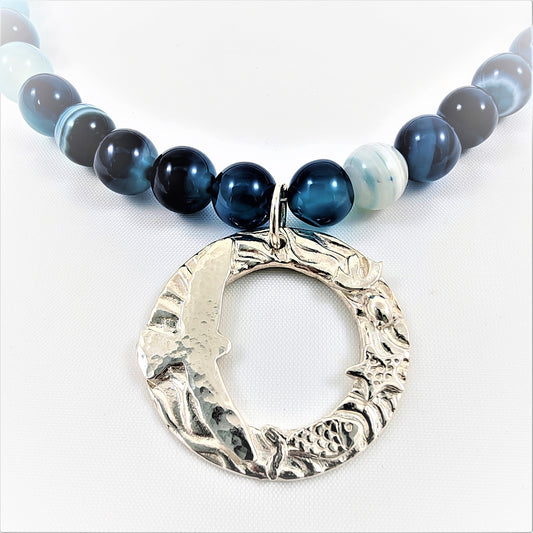 This exquisite necklace features a round pendant depicting a seagull over the sea, accompanied by fish and starfish in the rippling waters. Adjustable in length, it is enhanced with a vivid blue beaded agate necklace. The pendant is 999 silver the attachments 925 sterling silver.