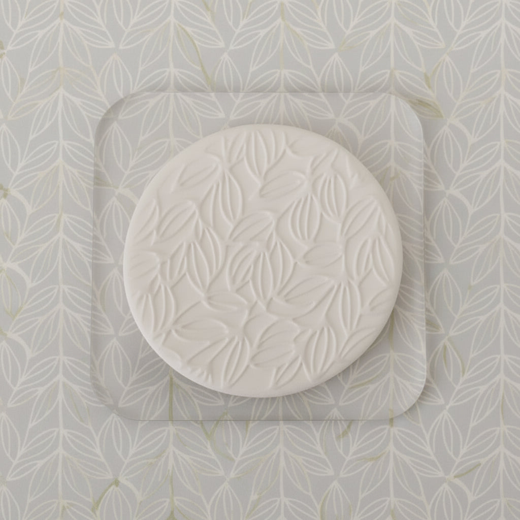 A round stamp with an embossed leaf pattern, resting on a white square.