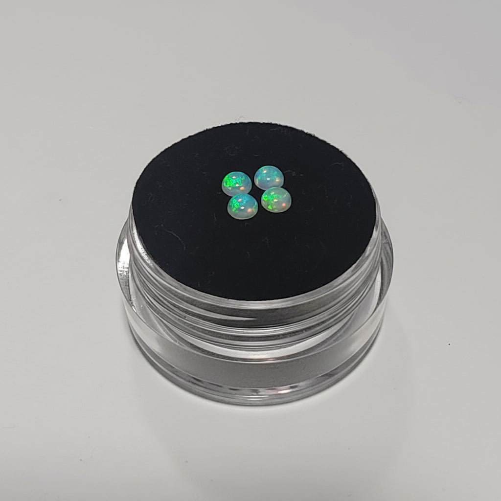 Three small iridescent cabochon gemstones with a blue-white base and green-orange flashes, displayed on a black circular pad atop a clear acrylic jar against a light gray background.